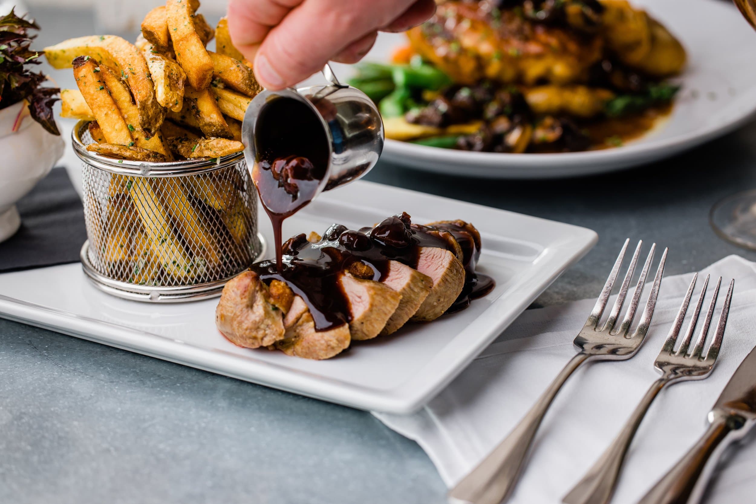 A hand pours sauce over sliced meat on a white plate next to a basket of fries.