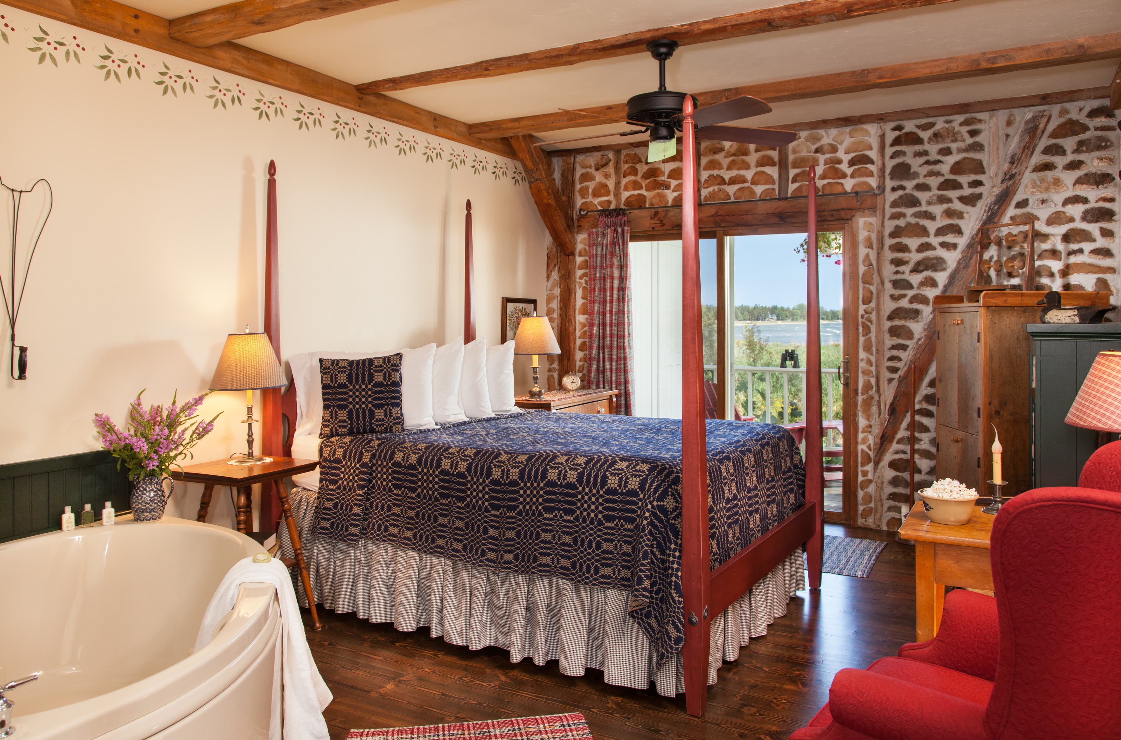 A rustic bedroom featuring a four-poster wooden bed with a blue patterned quilt, a stone accent wall, exposed wooden beams, and a sliding glass door opening to a balcony with a scenic view.