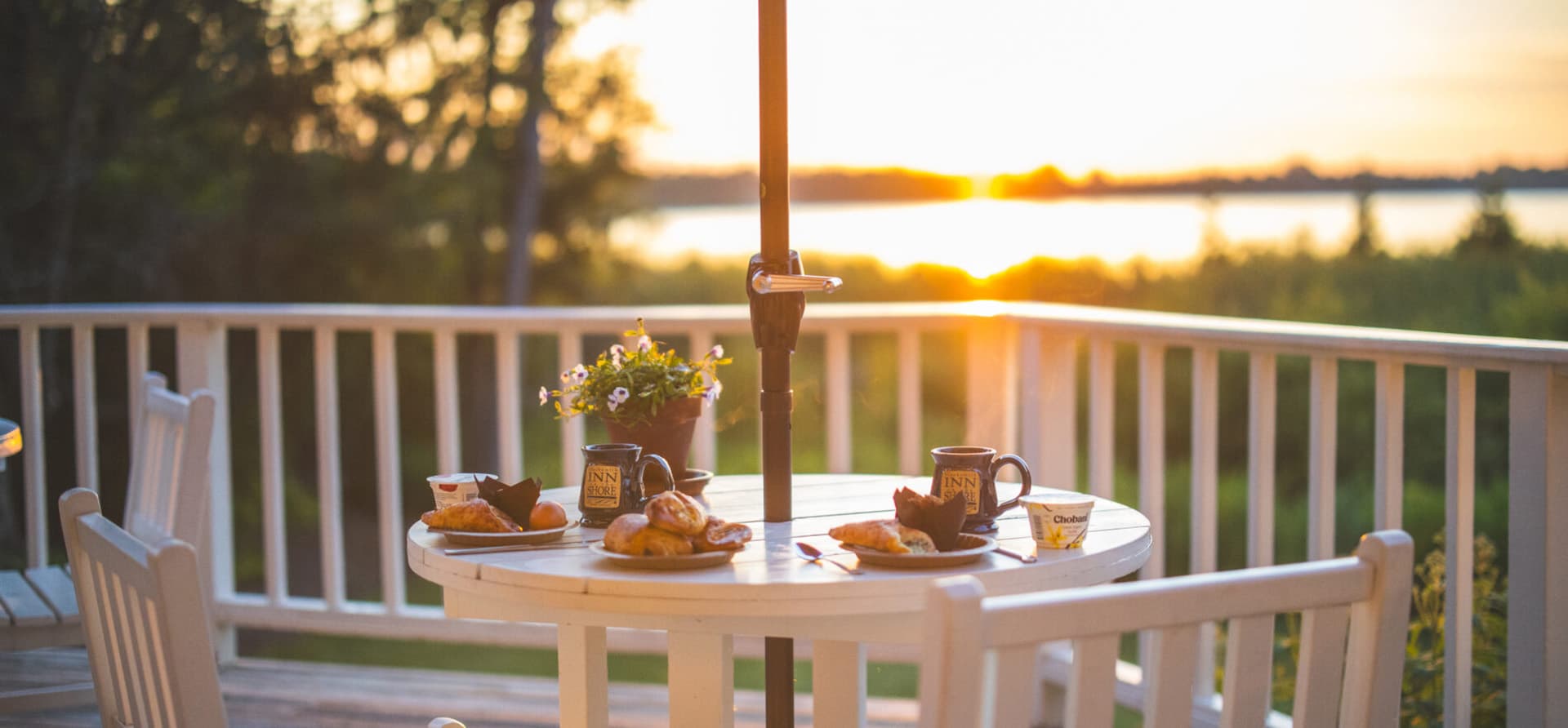 A well-set table with breakfast items and coffee mugs overlooks a serene sunset by the water.