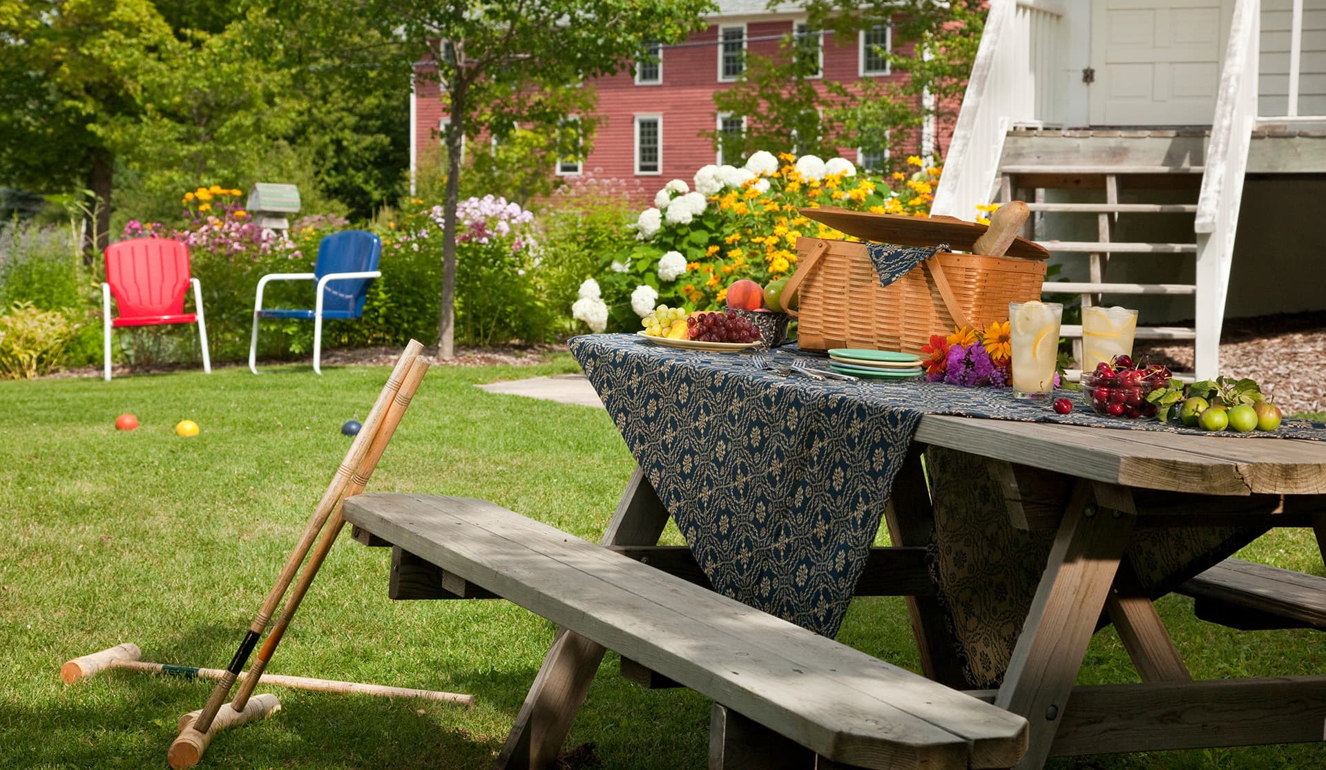 A picnic setup with food and drinks on a table in a garden, surrounded by colorful flowers and chairs.