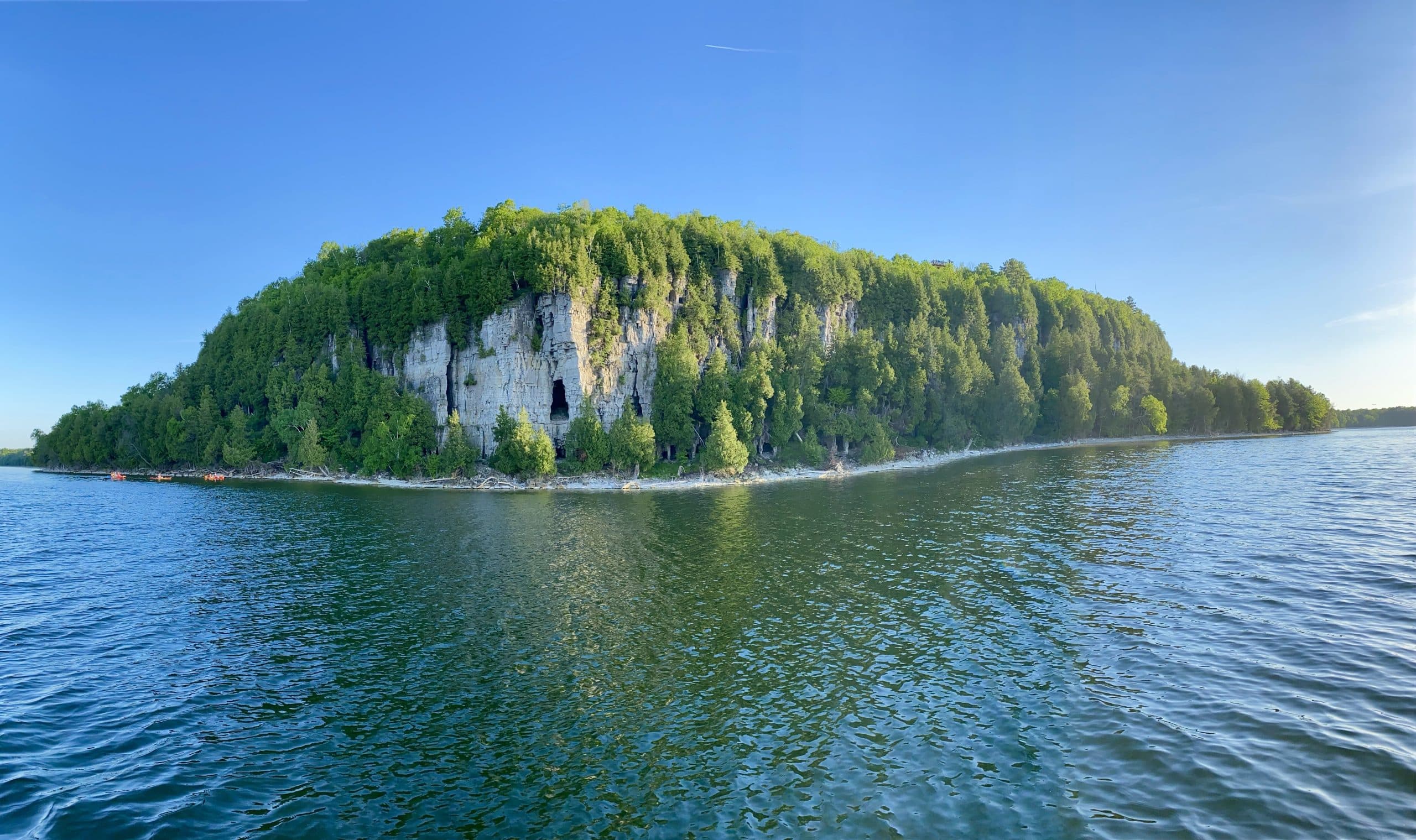 A panoramic view of a lush, green cliff rising above calm blue waters under a clear sky. A panoramic view of a lush, green cliff rising above calm blue waters under a clear sky.
