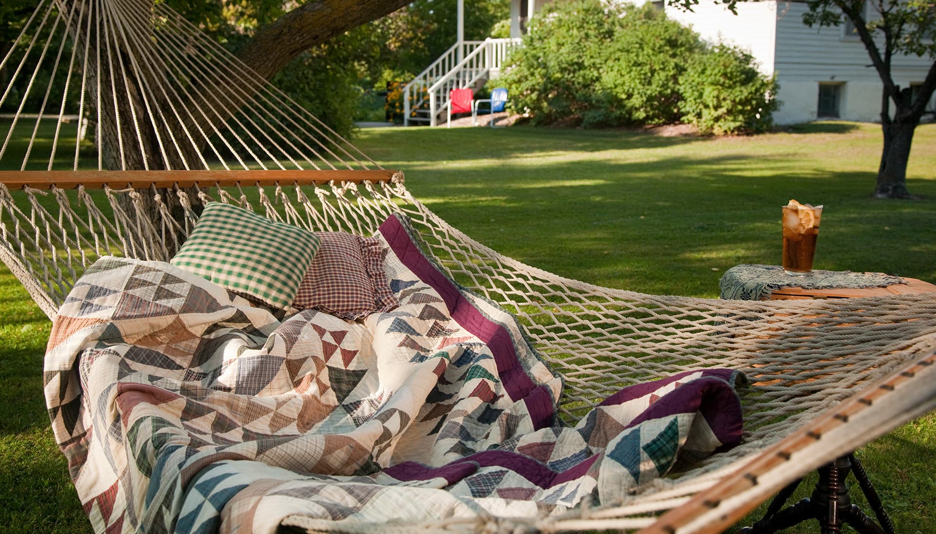 A cozy hammock with a quilt and pillows is set in a sunny yard beside a drink on a side table.