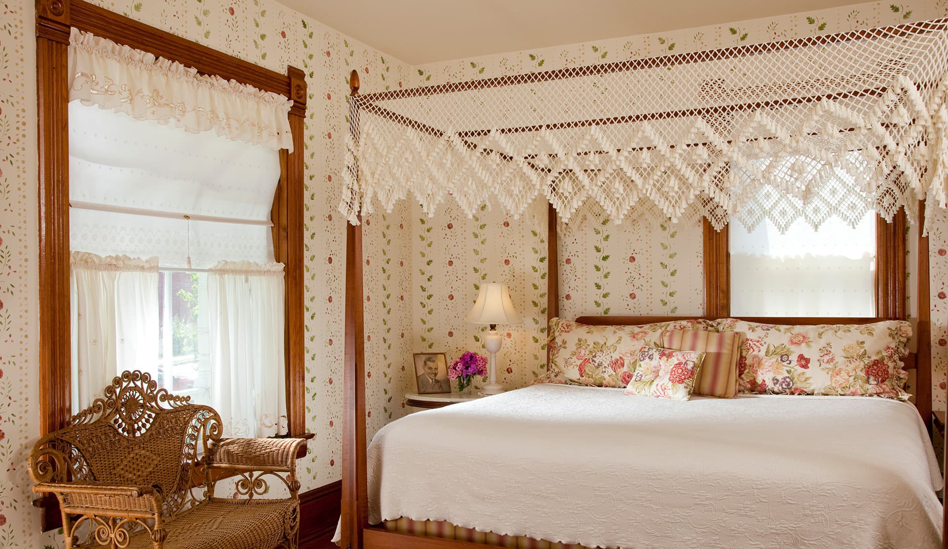 A cozy bedroom featuring a canopy bed, floral wallpaper, and a vintage chair.