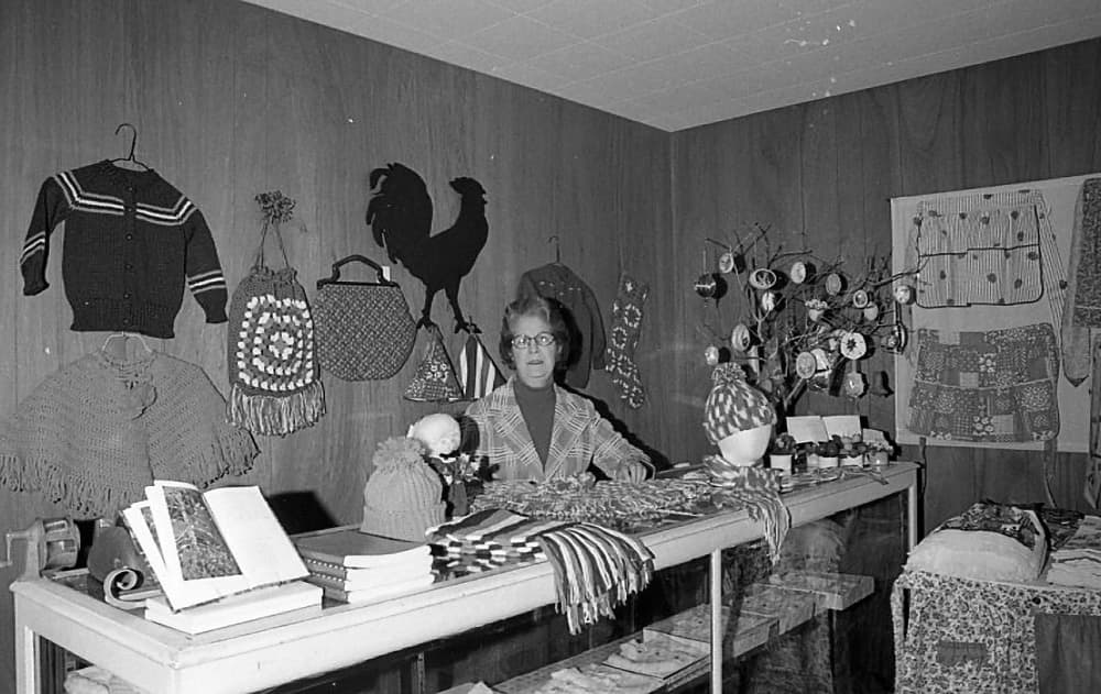 A woman sits behind a display of handmade crafts in a vintage shop, surrounded by knitted items and decorative accessories.